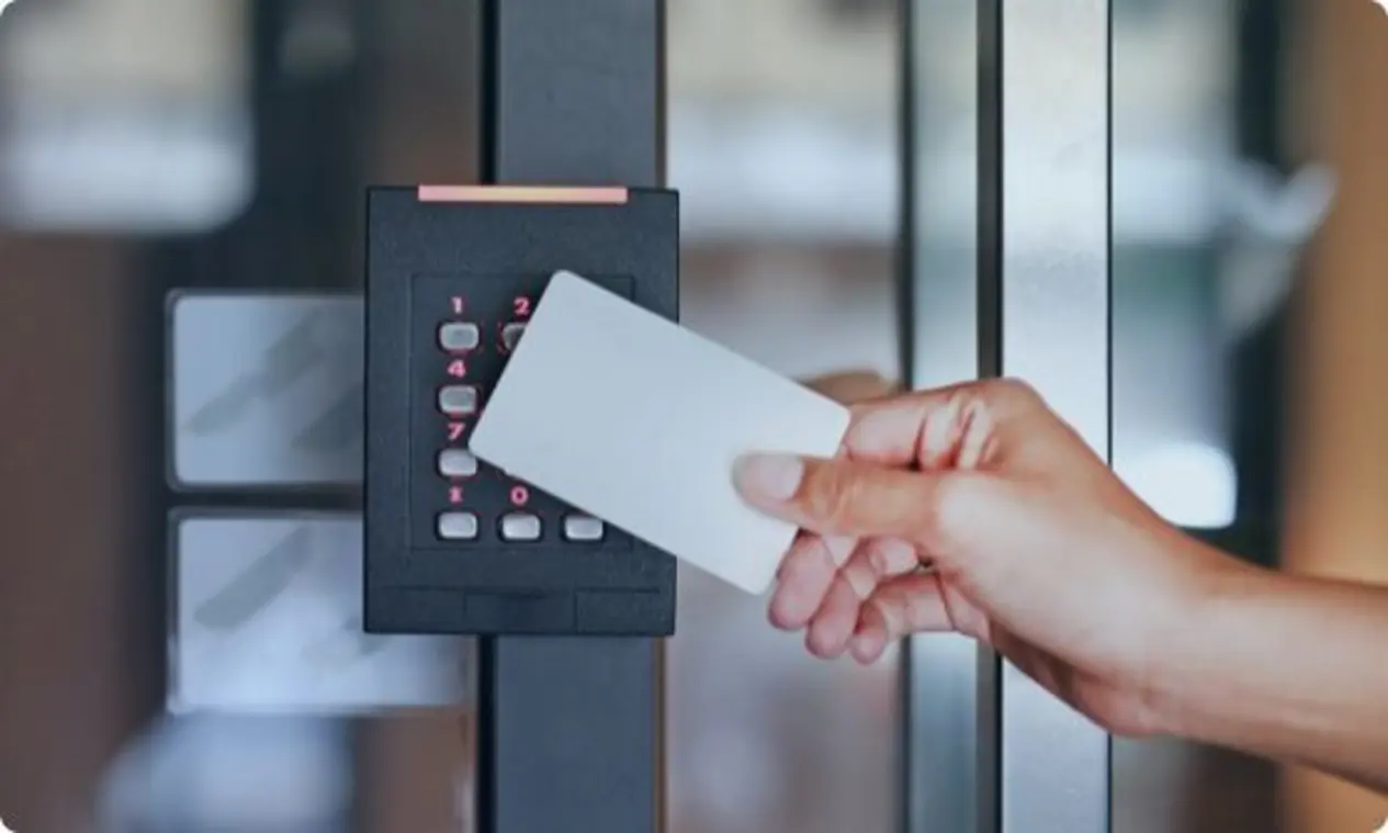 A hand holding a white card near a keypad access control system mounted on a door or panel. The keypad has numbers and buttons, suggesting it's used for entering a code to access a secured area indoors.