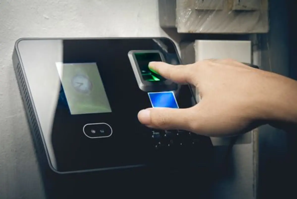 A biometric fingerprint scanner with a hand positioned to scan a fingerprint. The device features a digital display showing a clock and possibly other details, likely used as a security or access control system for identity verification.