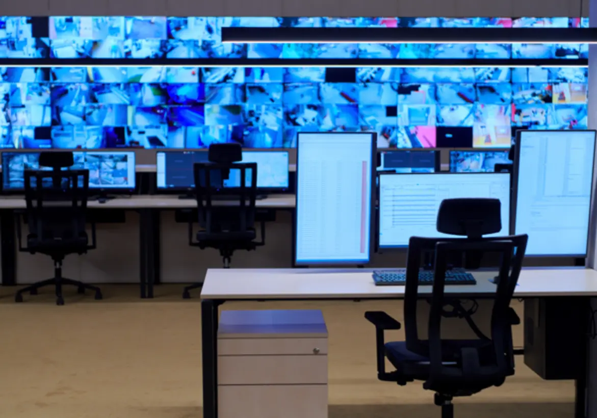 A control room featuring multiple workstations with computers and large screens displaying numerous video feeds, likely from security cameras. The desks are equipped with monitors and office chairs, and a filing cabinet is visible in the foreground, indicating a focus on surveillance or monitoring activities.