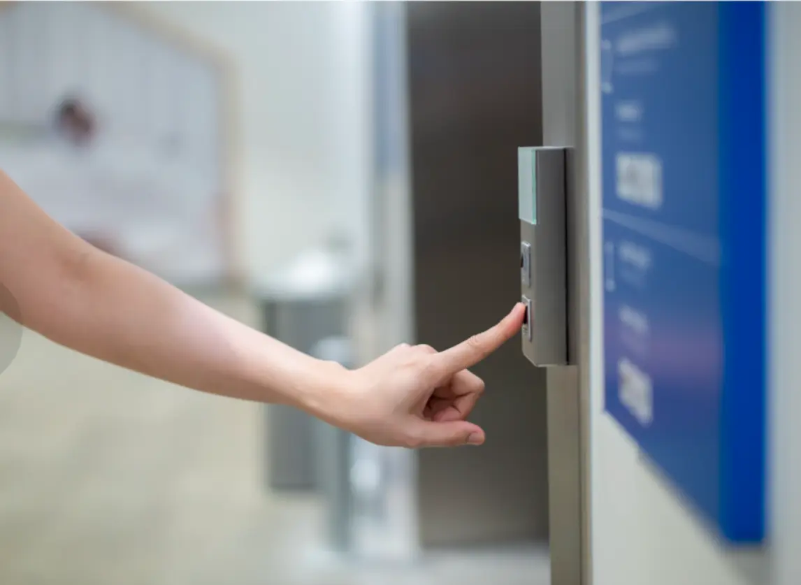 A close-up of a hand pressing a button on a control panel, possibly for an elevator or access system, with a softly blurred hallway or lobby visible in the background.