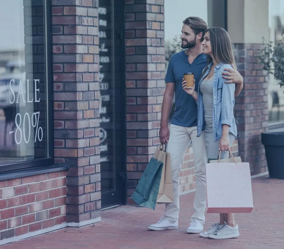 A smiling young couple stands on a brick-paved sidewalk in front of a retail store with a large "SALE -80%" sign in the window. The man, wearing a blue t-shirt and light pants, holds several shopping bags and a coffee cup while looking at his partner. The woman, in a striped dress and denim shirt, holds a large white shopping bag and smiles back at him. His arm is gently around her shoulder, capturing a joyful moment of retail therapy and togetherness.