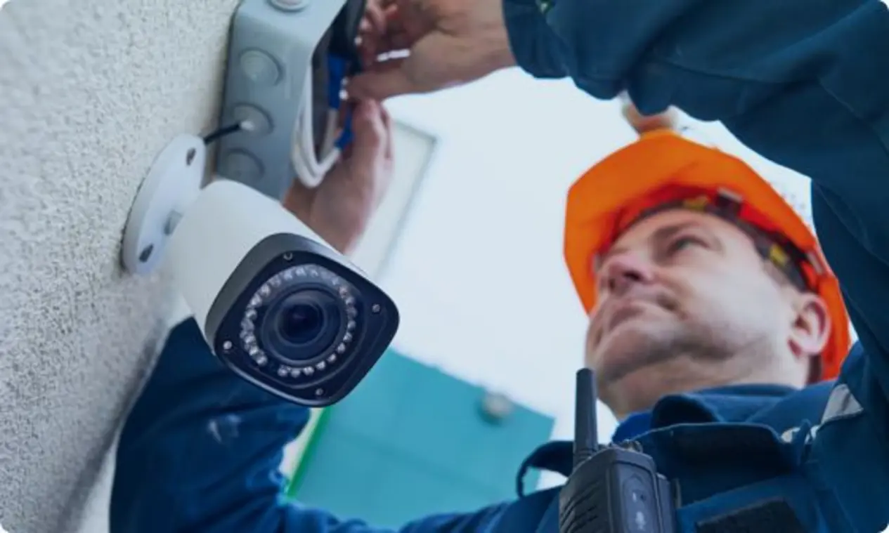 A person wearing an orange safety helmet and blue work uniform works on installing a surveillance camera mounted on a wall. Their hands are connecting wires or components related to the camera. A walkie-talkie is visible on their chest, emphasizing a focus on communication and safety.