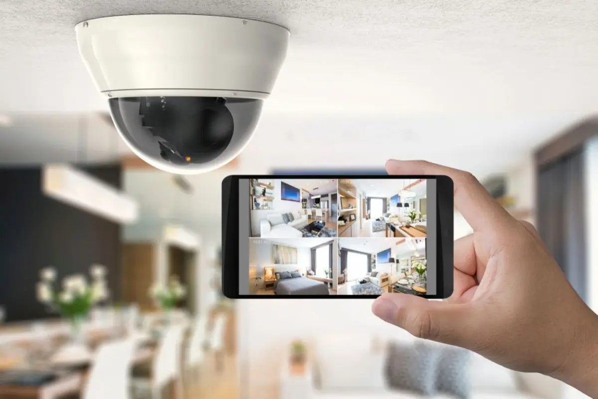 A security camera mounted on the ceiling, with a person holding a smartphone displaying multiple camera views of a home interior. The background shows a well-lit living or dining area, illustrating a security or monitoring system in use.