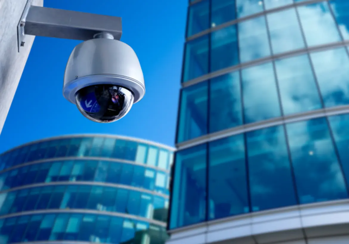 A dome-style security camera is mounted on the exterior of a building. In the background, large reflective glass structures with modern architectural design can be seen, mirroring the sky.