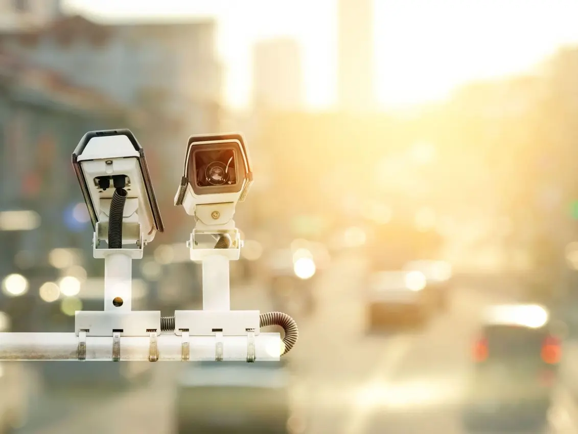 Two security cameras mounted on a pole overlook a street scene with blurred traffic and urban structures in the background. The warm, glowing light suggests it's either sunrise or sunset, creating a serene yet watchful atmosphere.