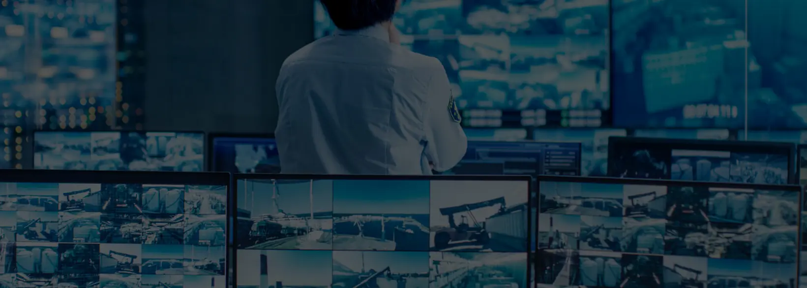 A person sitting in a high-tech control room, surrounded by multiple computer screens displaying surveillance footage from various locations, suggesting a focus on security monitoring and data analysis.