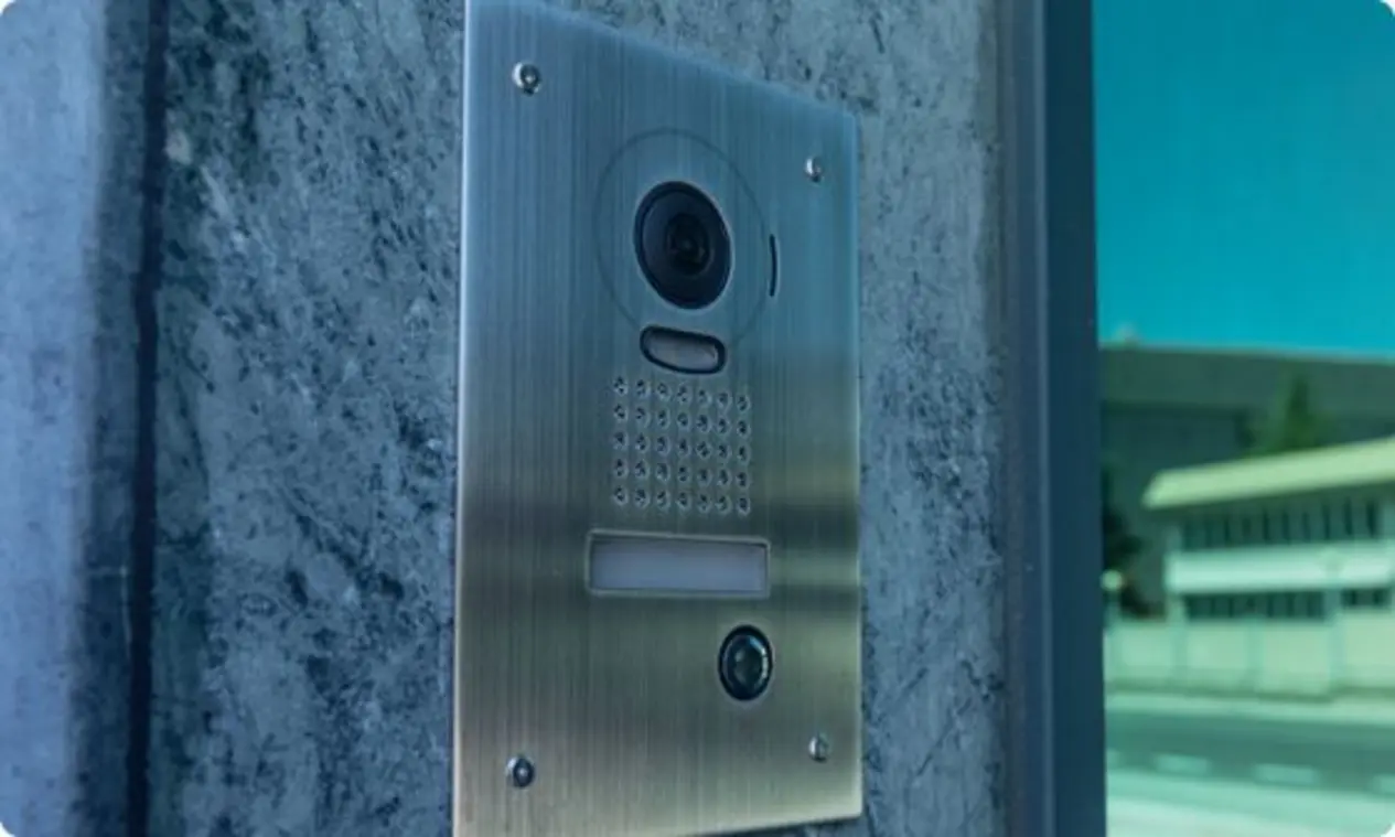 A metallic intercom or doorbell system mounted on a wall, featuring a video camera, perforated speaker area, and a communication button. Designed for entry points, it enables communication between visitors and occupants inside a building.